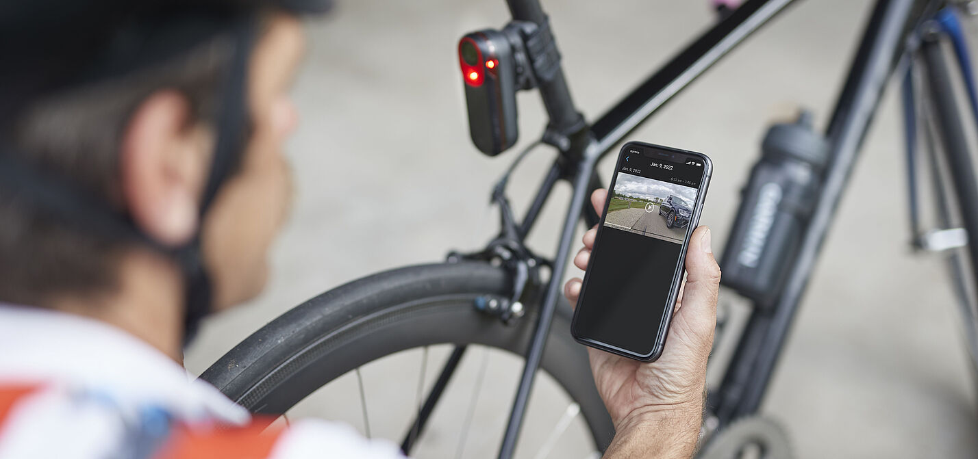 Rücklicht Garmin Varia RCT715 Mann kniet mit Smartphone in der Hand vor einen Rennrad mit rot leuchtendem Rücklicht an Sattelstütze. Das Rücklicht Garmin Varia RCT715 mit integrierter Kamera zeichnet permanent auf und speichert Daten aber nur bei einem erkannten Unfall.